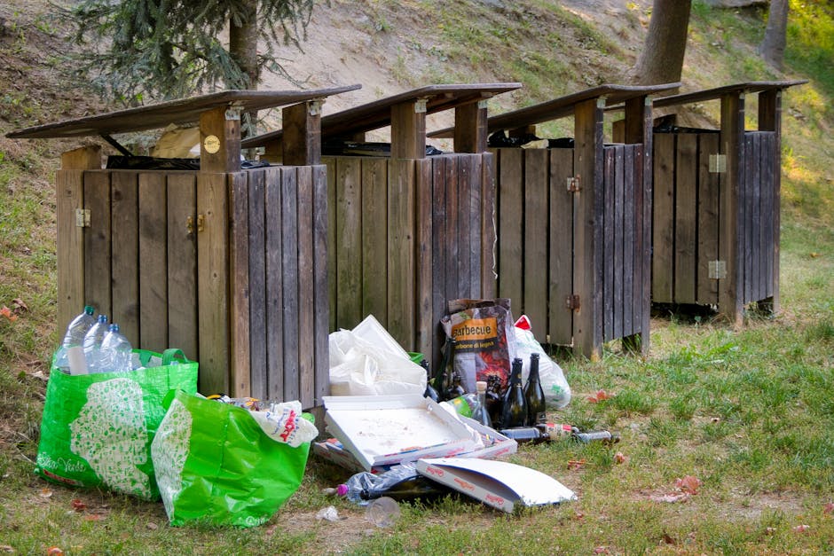 A row of five large wheelie bins arranged along a paved pathway beside a brick wall, with each bin having a distinct colour and lid design. Starting from the front, there is a green bin with a ribbed